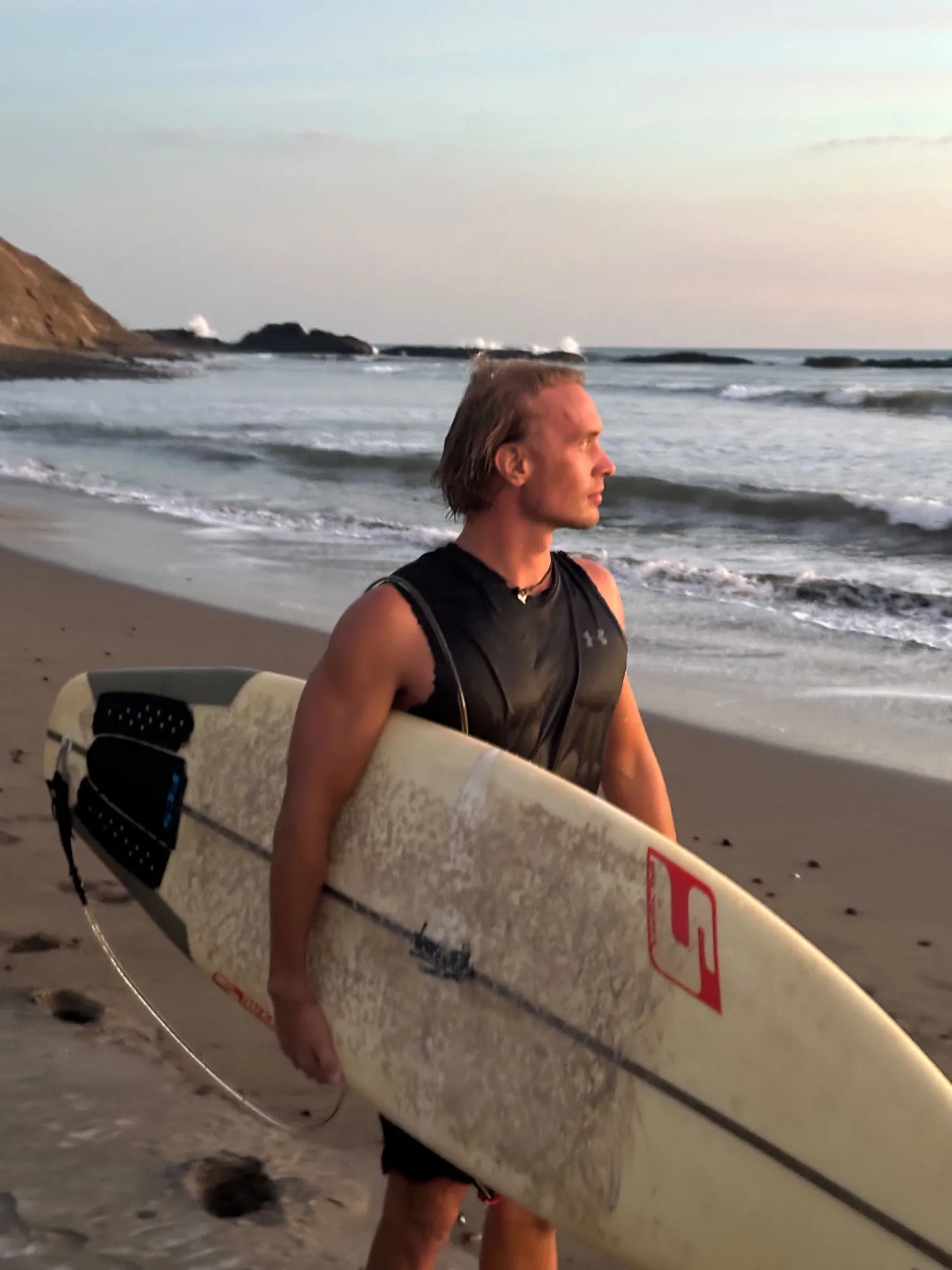 The founder of Pelagos on the beach at sunset, holding a surfboard and looking out at the waves